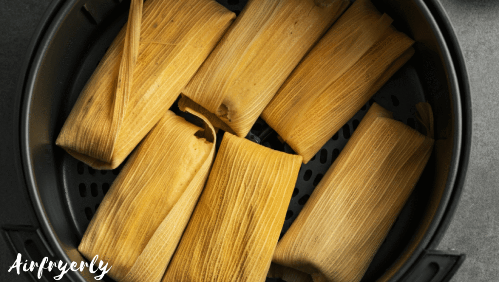 Frozen tamales arranged in a single layer inside an air fryer basket with husks still on