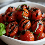"Air fryer cherry tomatoes served in a rustic white bowl, roasted to perfection with caramelized edges, herbs, and balsamic glaze, placed on a linen cloth with basil and tomato stems around."