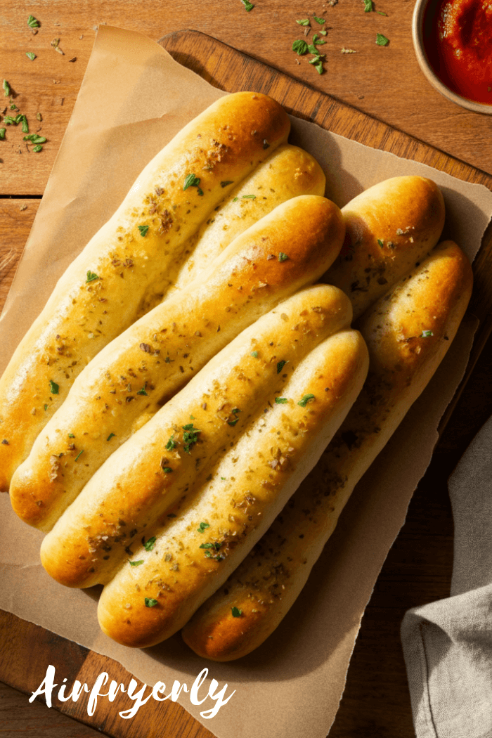 Golden New York Bakery breadsticks served on a white plate with dipping sauces, garnished with fresh parsley and placed on a rustic wooden table beside an air fryer basket